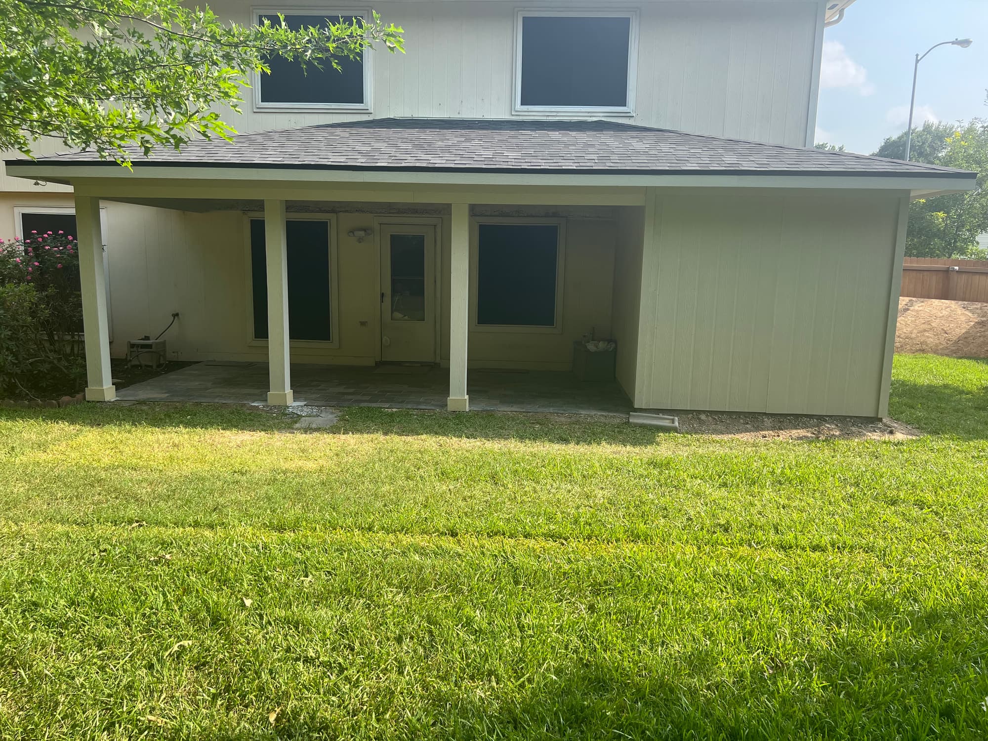 Completed covered patio addition with shingled hip roof, painted posts and a shaded paver floor