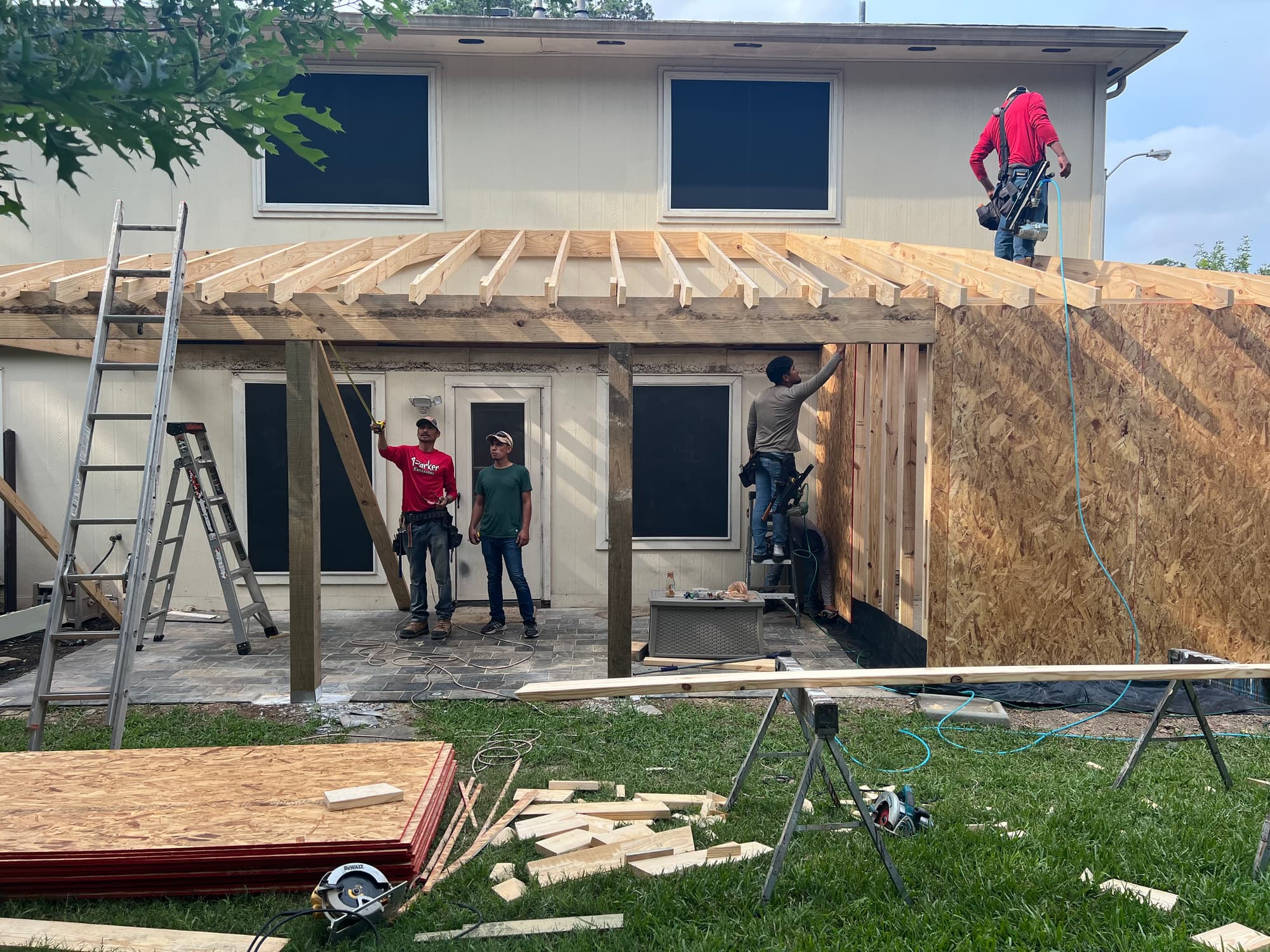 Three crew members framing the rafters and sheathing the wall of a new covered patio addition behind a two-storey house